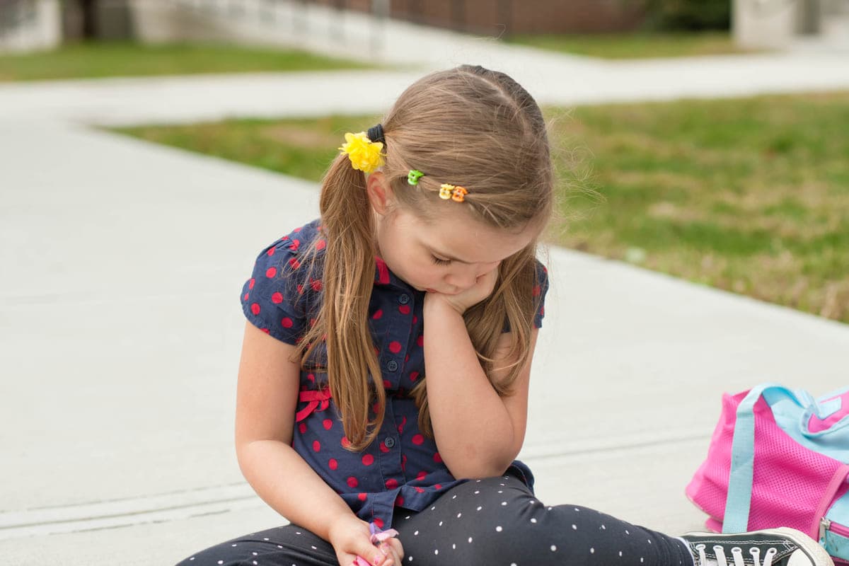 Very sad little girl sitting on a sidewalk in front of school.