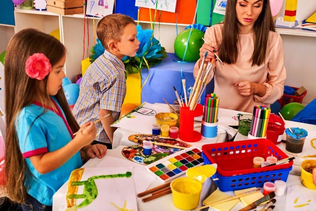 Children painting and drawing in kids club. Craft lesson in primary school. Kindergarten teacher and small students work together. Kid boy and girl coloring picture in class. Mess in classroom.