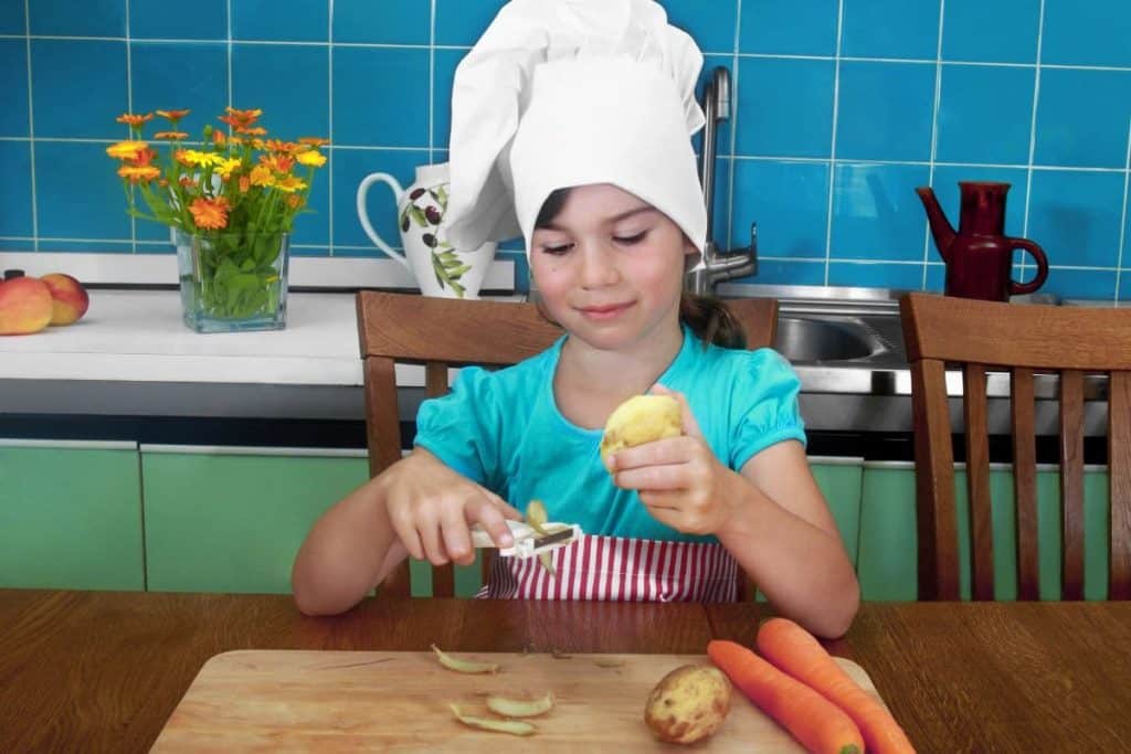 Girl in chef hat peels potatoes and carrots in kitchen.