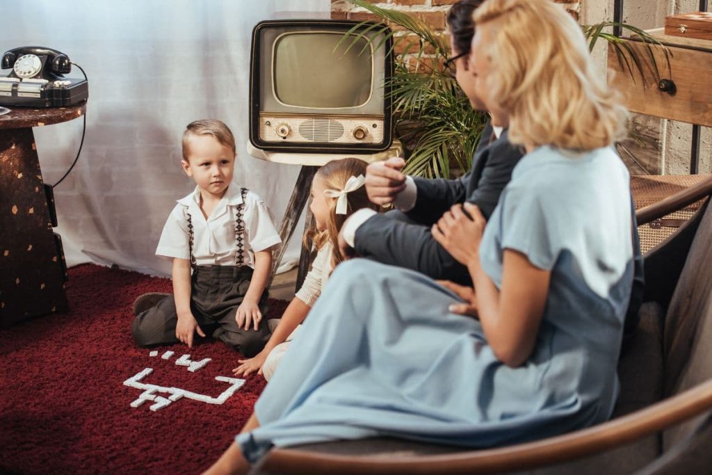 Parents sitting on sofa and looking at adorable children playing at home with vintage theme.