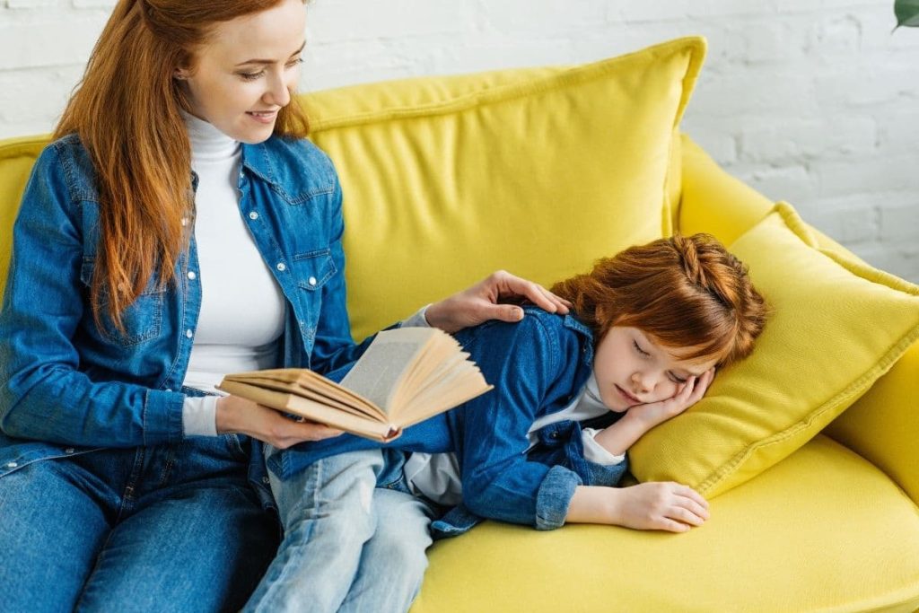 a mother reading a book to her sleepy kid on a sofa.