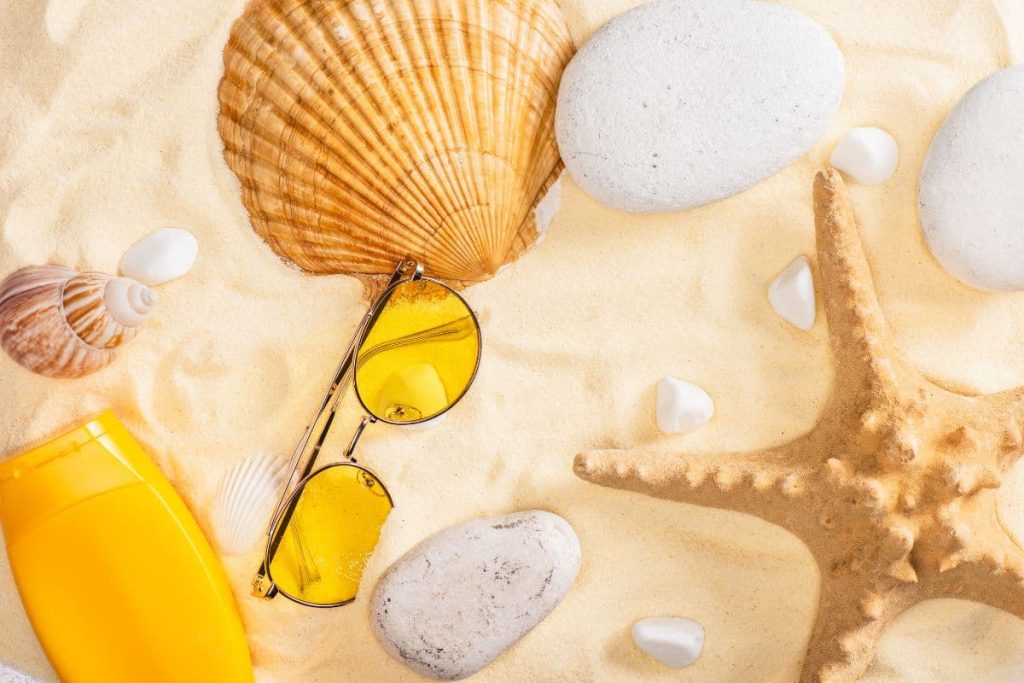 a sunglassess with lotion essentials and sea shells on a sand beach.
