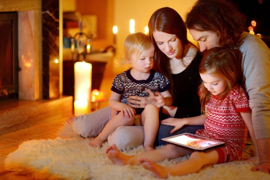 Happy young family using a tablet pc at home by a fireplace in warm and cozy living room on winter day.