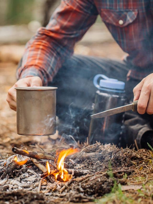 man traveler hands holding mug with water near the fire outdoors. bushcraft, adventure, travel, tourism and camping concept.