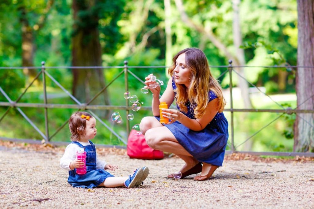 Happy young mother and adorable toddler girl blowing soap bubbles and having fun together, outdoors. Woman and daughter playing on warm sunny day in park.